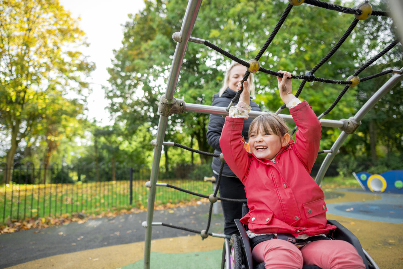 A cheerful low angle medium close-up of a young girl who is a wheelchair user playing in the park on the climbing frame with her mother. They're in a playground ina. public park in Newcastle upon Tyne in the North East of England