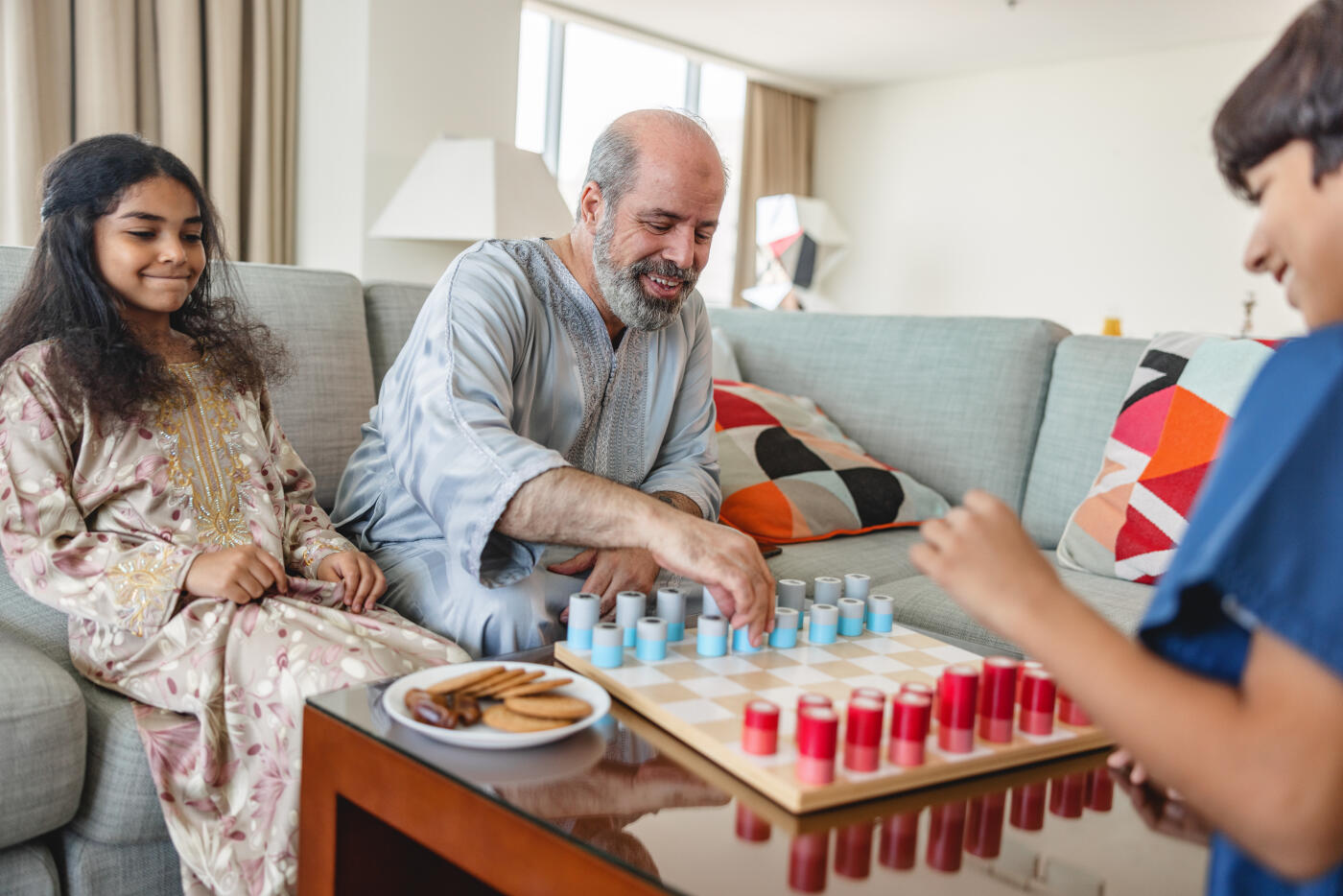 Middle eastern grandfather is enjoying spending time with his grandchildren. They are all smiling and wearing traditional clothes. They are sitting on large grey sofa in the living room. Beautiful interior design in the background.