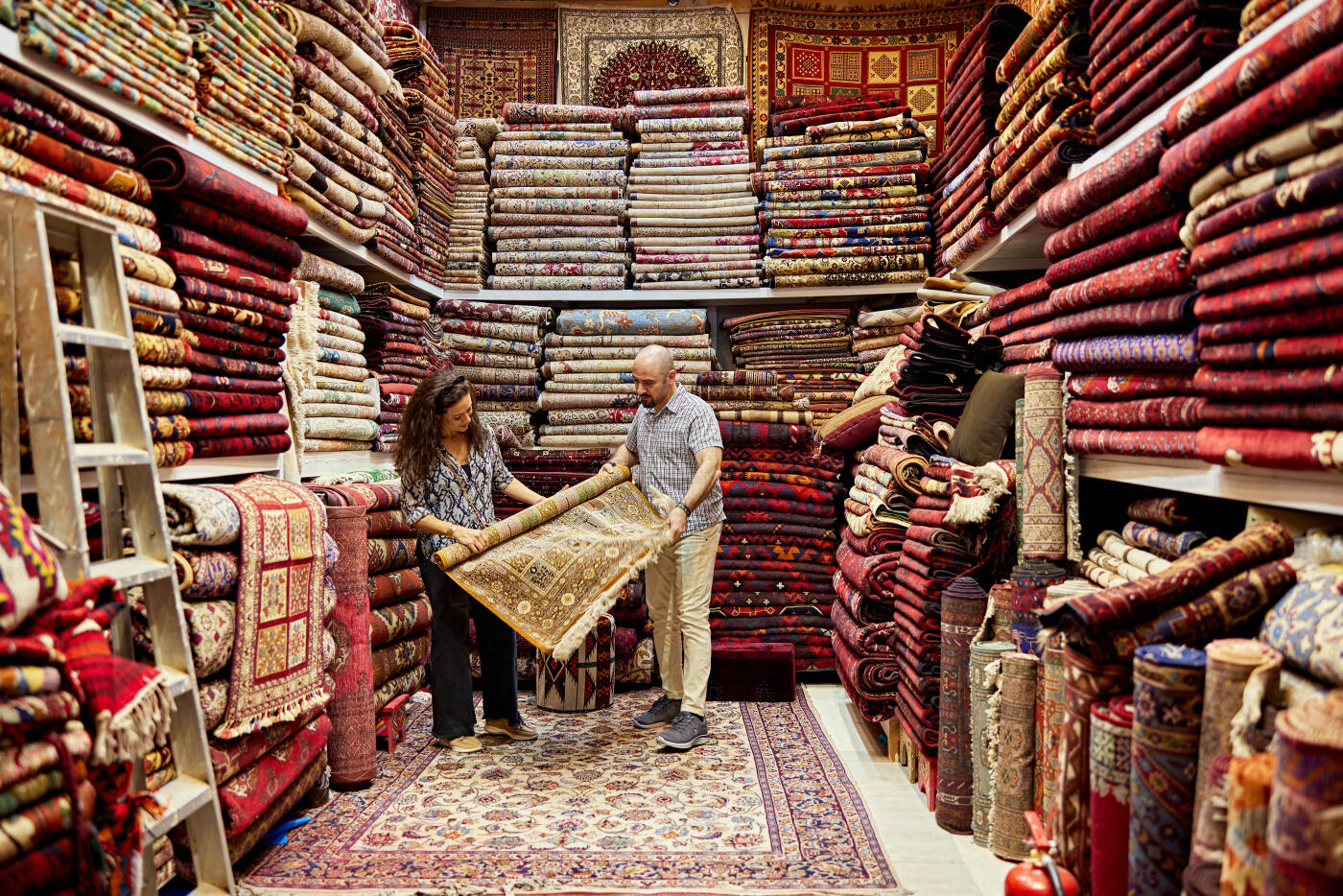 Full length view of couple in 30s and 40s examining one of many handwoven rugs from Saudi Arabia, Iran, Kashmir, and Afghanistan.