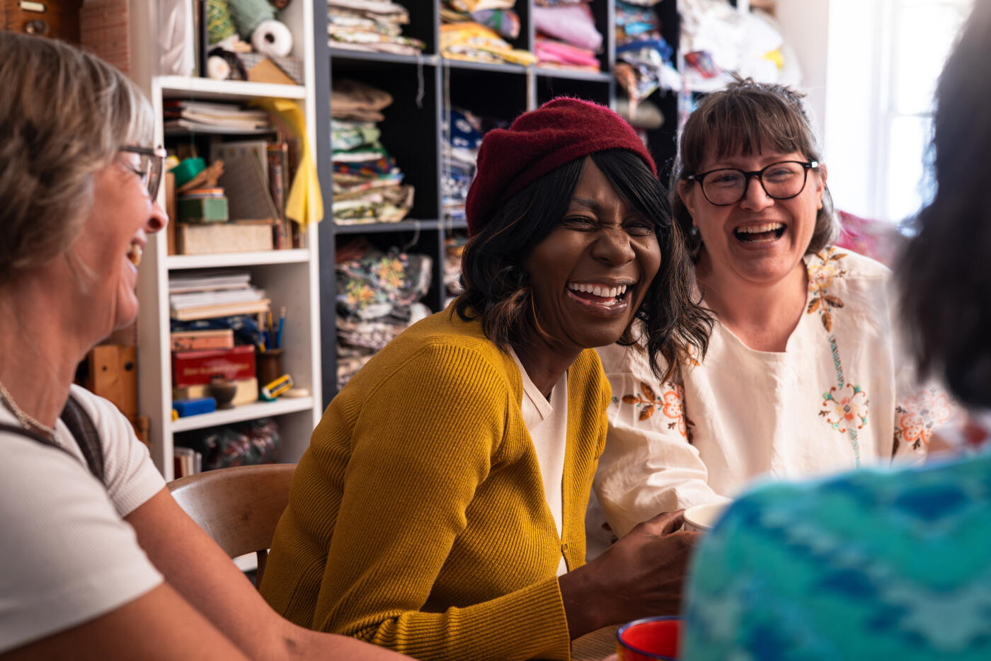 An over-the-shoulder view of a small business owner running a sewing club at her store, which upcycles fabric into products she sells. The club is all women, and they are sitting around a table enjoying cups of tea and cakes together before crafting.Videos are available similar to this scenario.