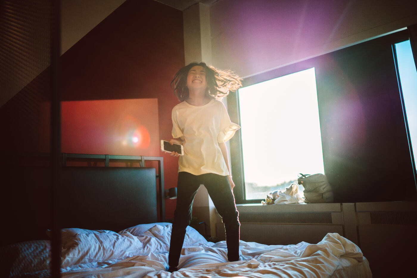 A joyful teenage girl jumps on a hotel bed while listening to music on her smart phone during family travel. Concept for emotional wellness, youth culture, and family vacation.