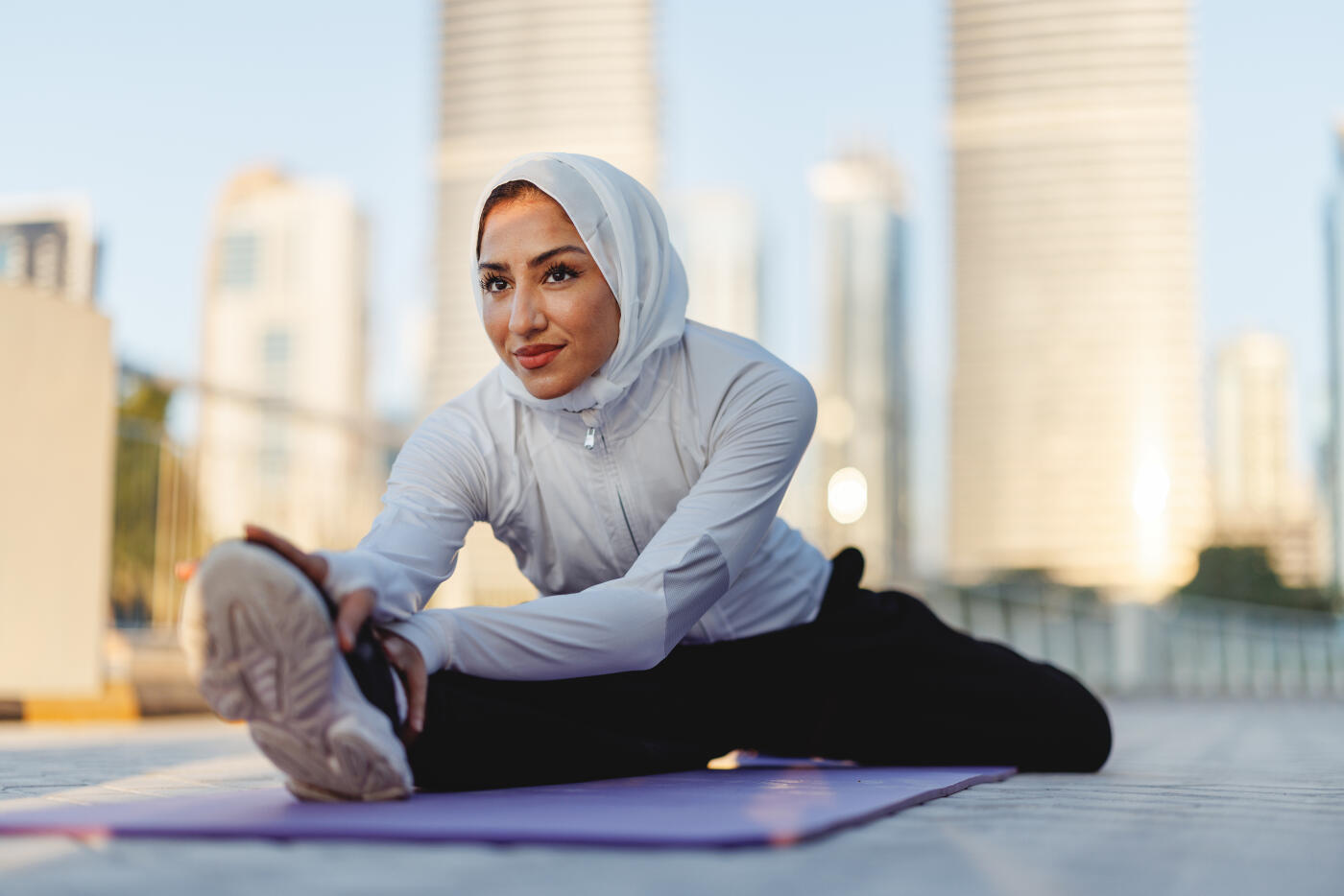A mid-adult mixed-race woman in a white hijab stretches on a yoga mat during a sunny morning in Dubai, with modern skyscrapers in the background, embodying focus and wellness.