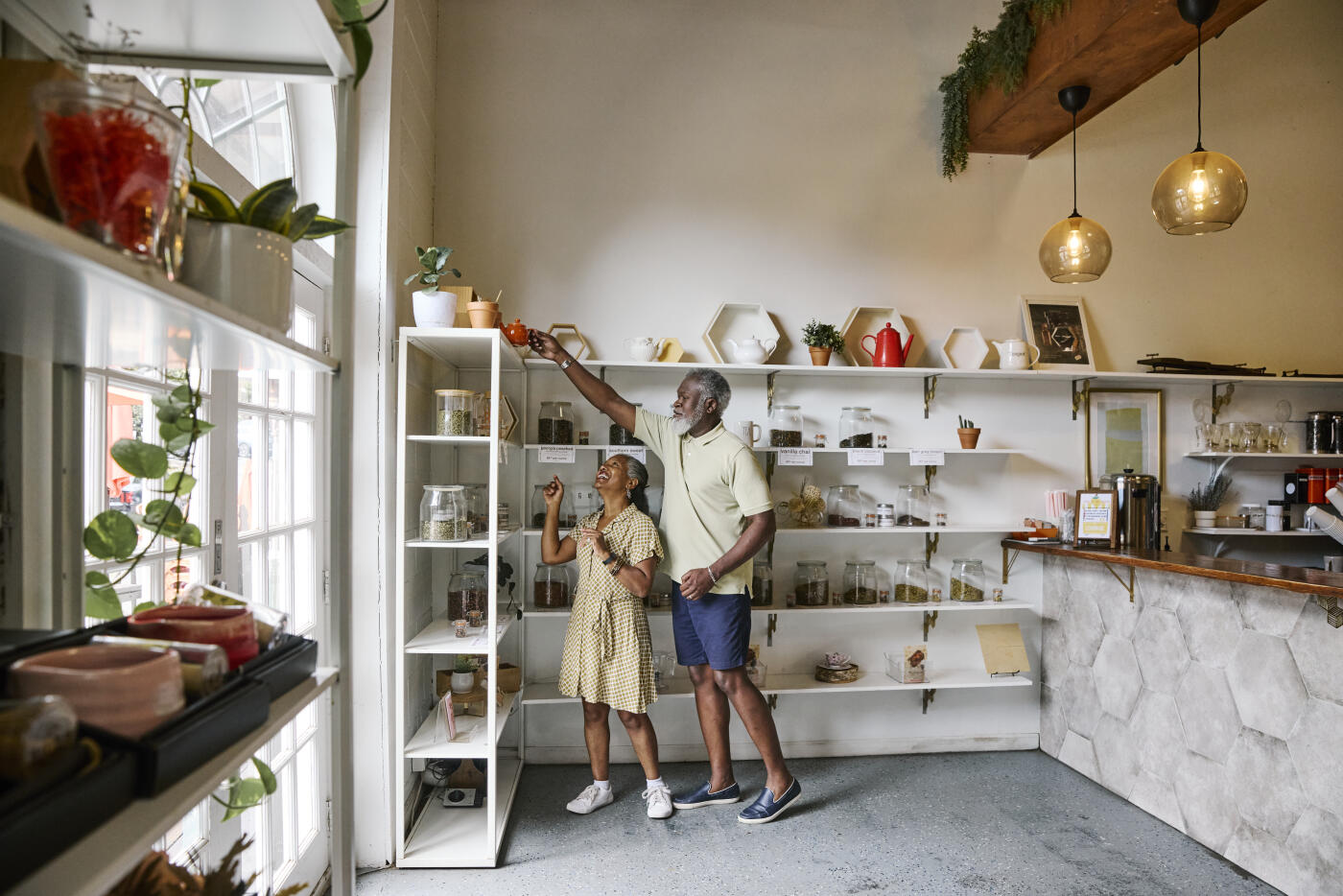 Mature man assisting a female friend in retrieving a teapot from a shelf in a retail setting.
