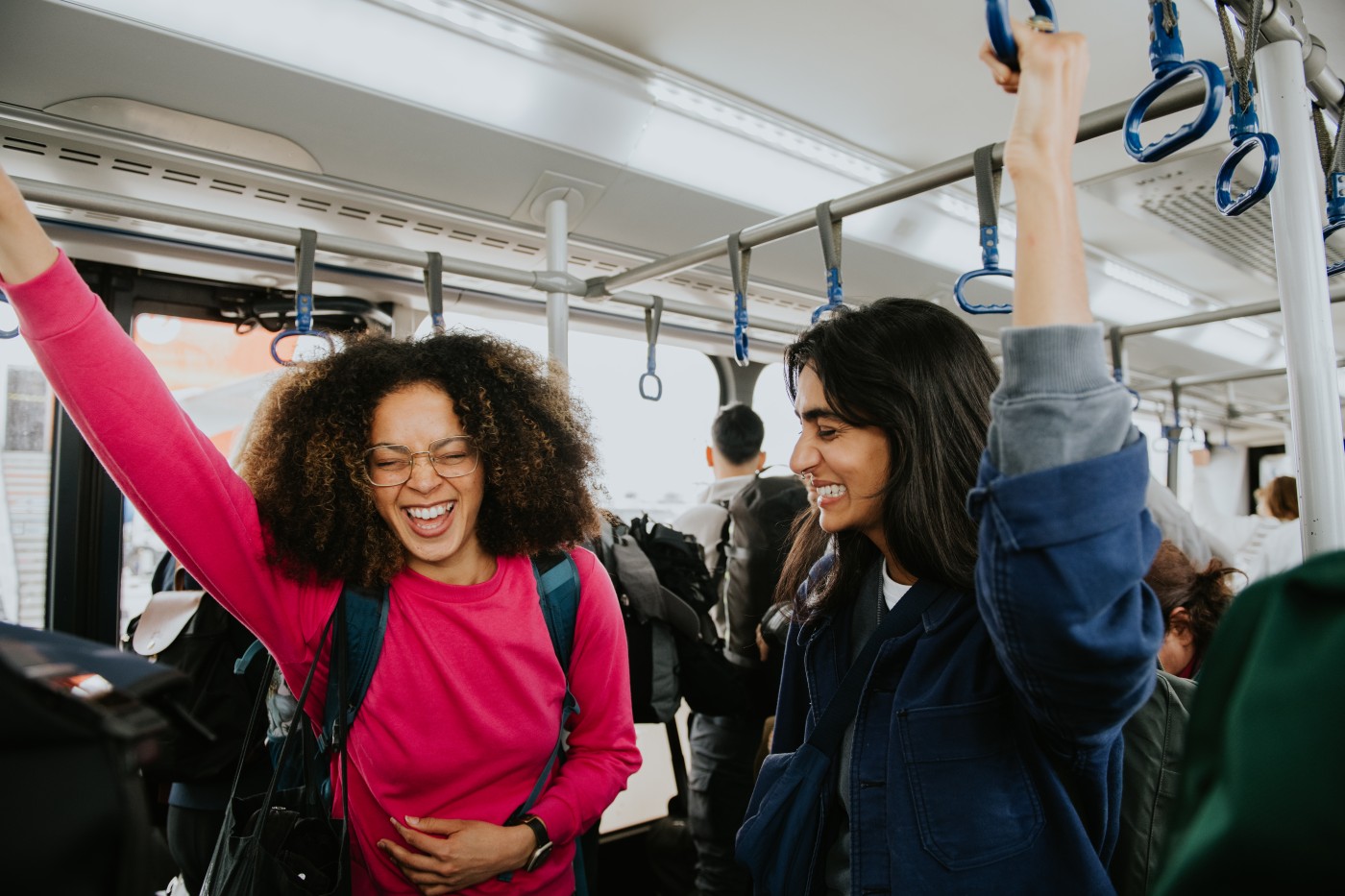 Two beautiful young woman standing in an airport transfer bus. They grip the handles on the ceiling to steady themselves. One of the woman is almost doubled over in laughter, as her friends giggles along at a joke they share.