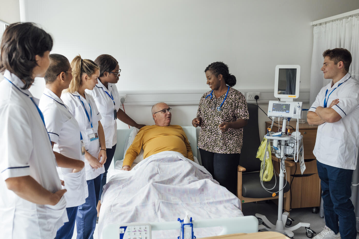 Group of medical students in a hospital standing around a male patient who is laying in a hospital bed. They are at a healthcare facility in Newcastle Upon Tyne, North East England. The students are all wearing their student nurse uniforms and are being taught by one lead doctor.