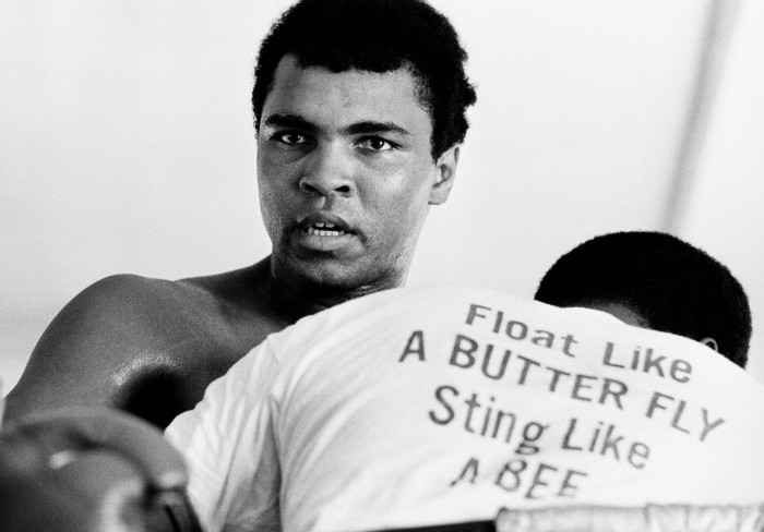American heavyweight boxing champion Muhammad Ali, circa 1970. The man in front of him is wearing a t-shirt printed with Ali's motto 'Float like a butterfly, sting like a bee'.