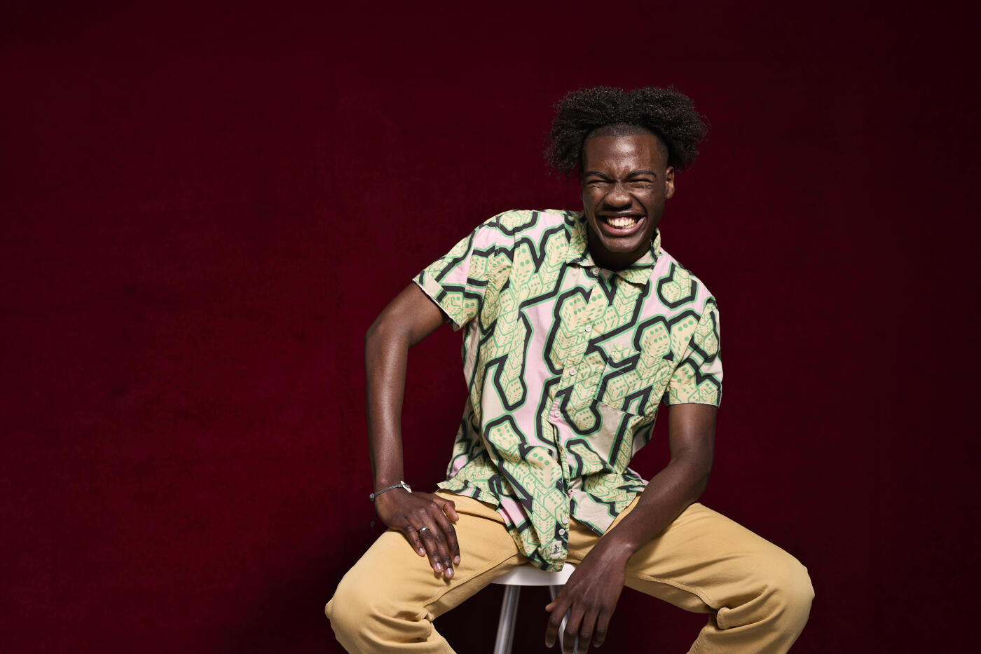 Portrait of young man with toothy smile sitting on stool against maroon background