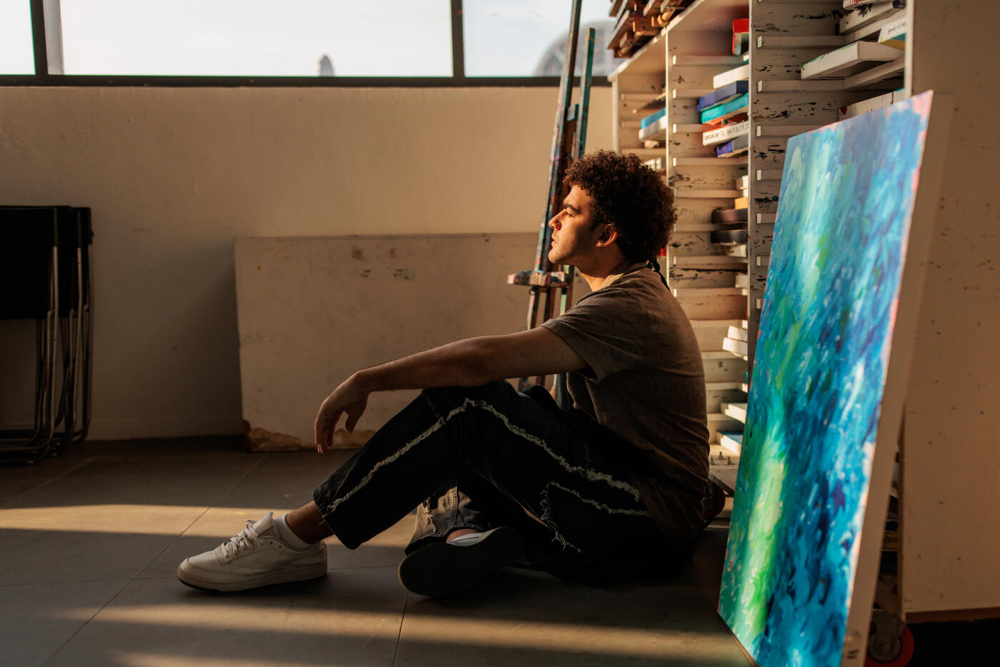 Male artist takes a break, sitting on the floor of his cozy workshop. A variety of her completed paintings are neatly arranged on shelves behind him, showcasing his creative journey.