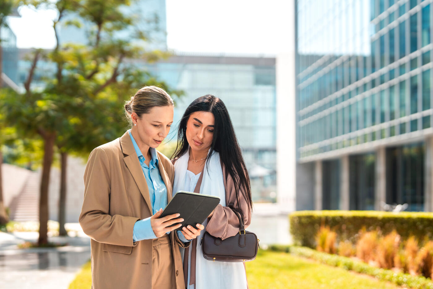 Two businesswomen examining a digital tablet together in an outdoor plaza surrounded by modern glass office buildings, green landscaping and natural daylight. Contemporary urban setting with professional attire and focused engagement.
