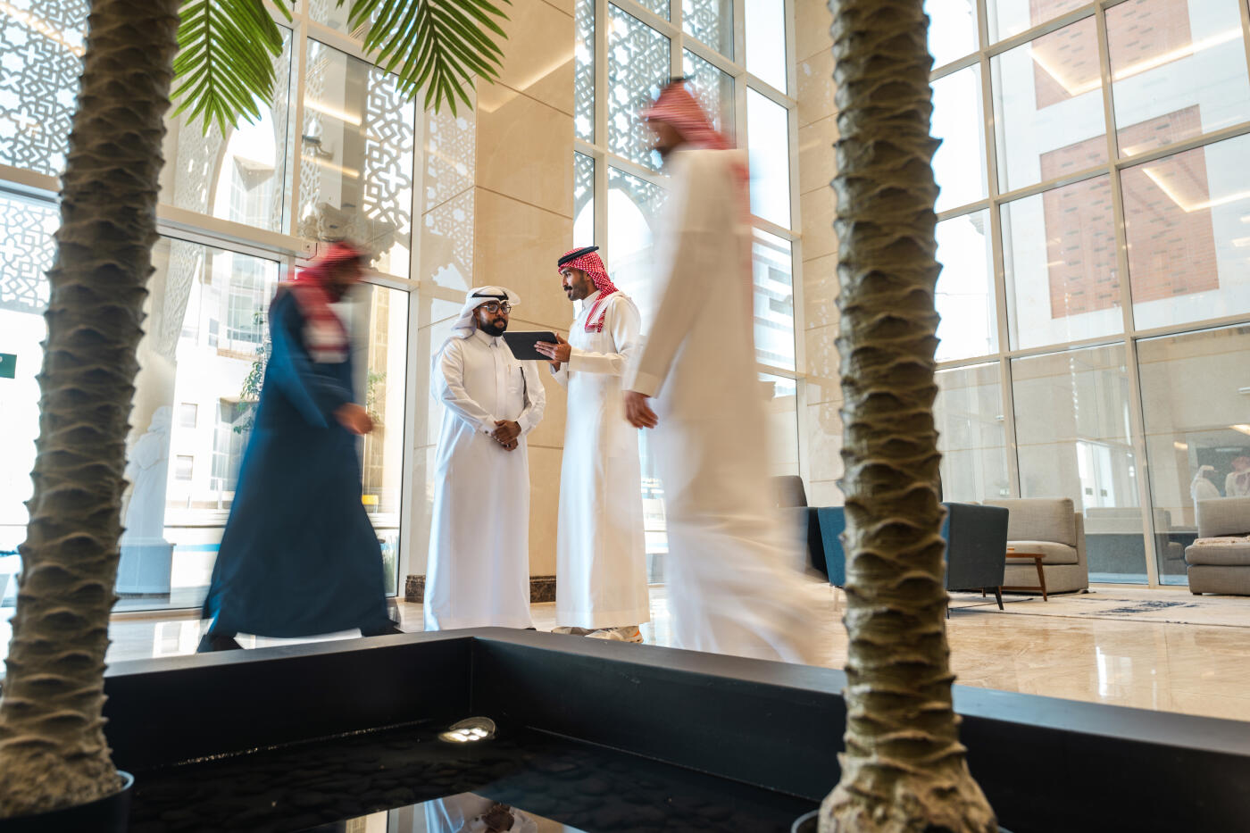 Saudi Arabianbusinessmen in modest clothing are seen collaborating in a contemporary hotel lobby, using a digital tablet. The space is bright with natural light, featuring modern furnishings and palm trees