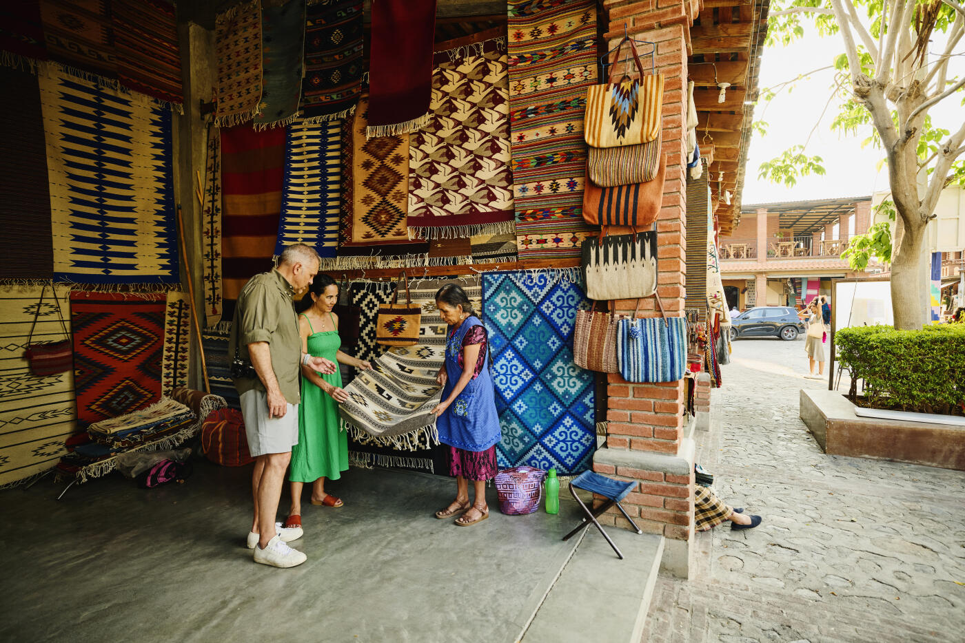 Wide shot of female local market vendor showcasing the patterns of a handcrafted rug to a curious senior couple in a Oaxacan market