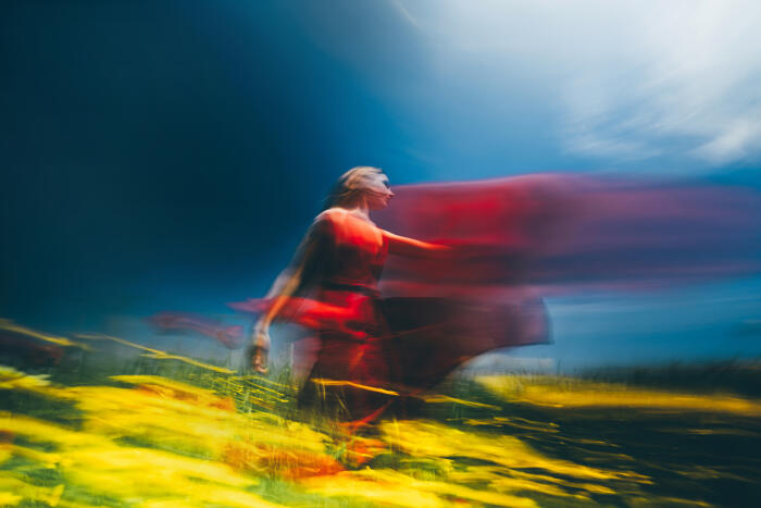 A young woman in a flowing red dress running through a field of yellow flowers under a dramatic sky in a dynamic motion blur.
