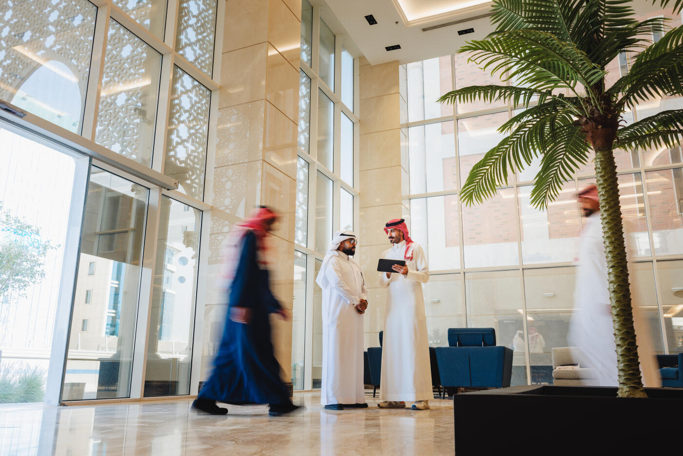 Middle Eastern businessmen in modest clothing collaborate using a digital tablet in a bright, modern hotel lobby. The space features large windows and a palm tree, creating a contemporary and professional atmosphere