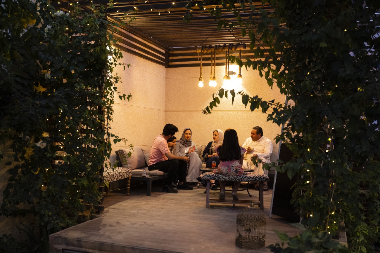 View through foliage and string lights of Middle Eastern children, parents, and grandparents spending weekend time together in secluded patio of Riyadh home.
