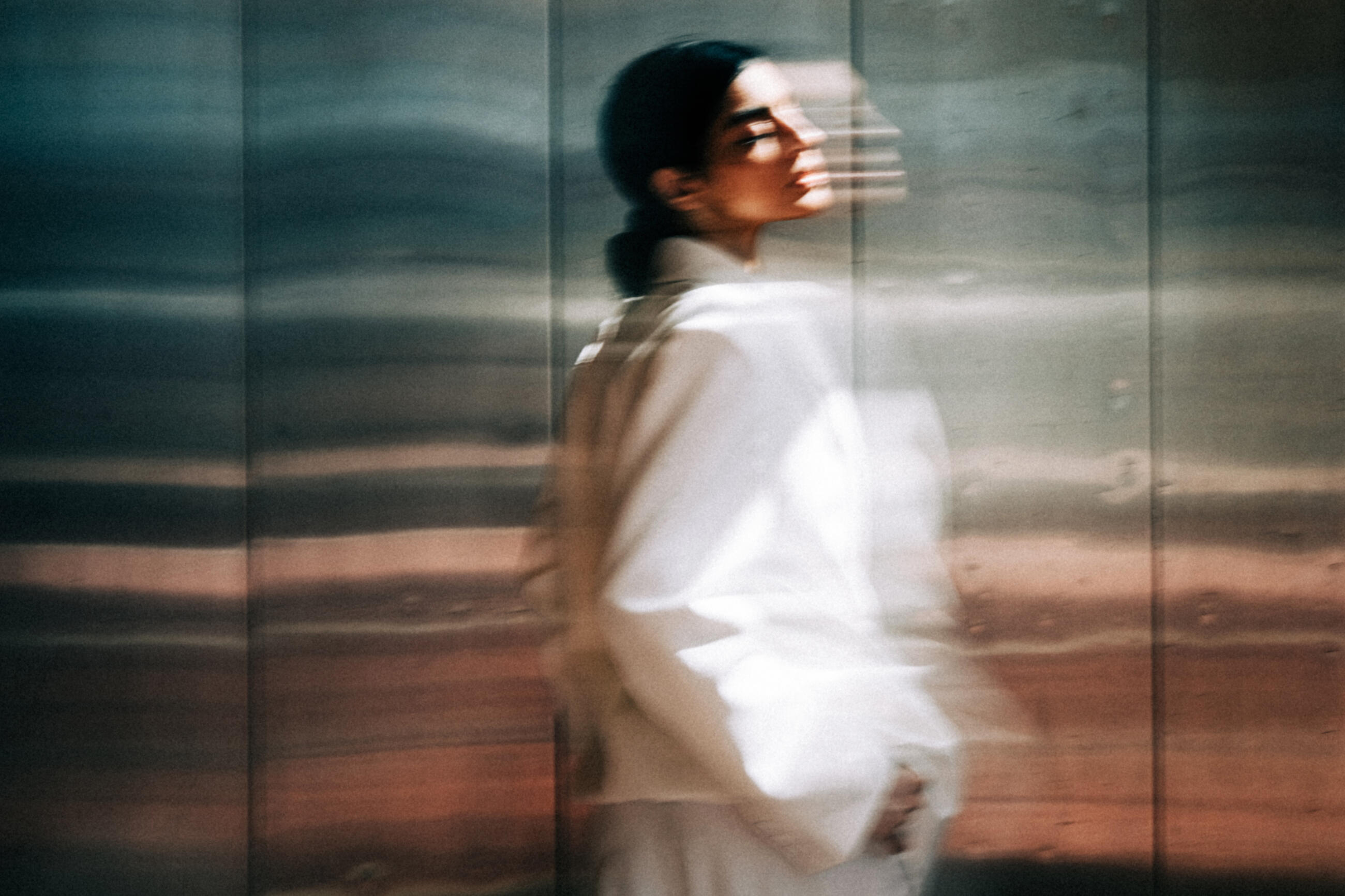A blurry portrait of a woman in a white jacket moving against a reflective metal wall.