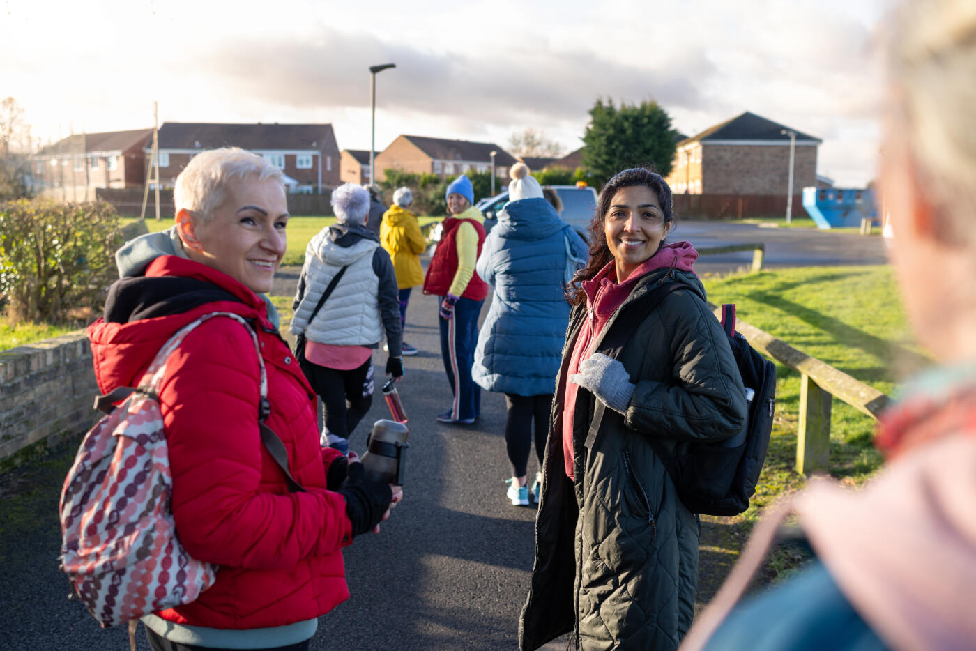 Over-the-shoulder shot of a group of mature women dressed in warm, sporty attire arriving to netball training together. They are smiling and carrying water bottles. The scene is located in a suburban park area, with houses and greenery in the background, illuminated by bright morning sunlight. 

Videos of this scenario are available