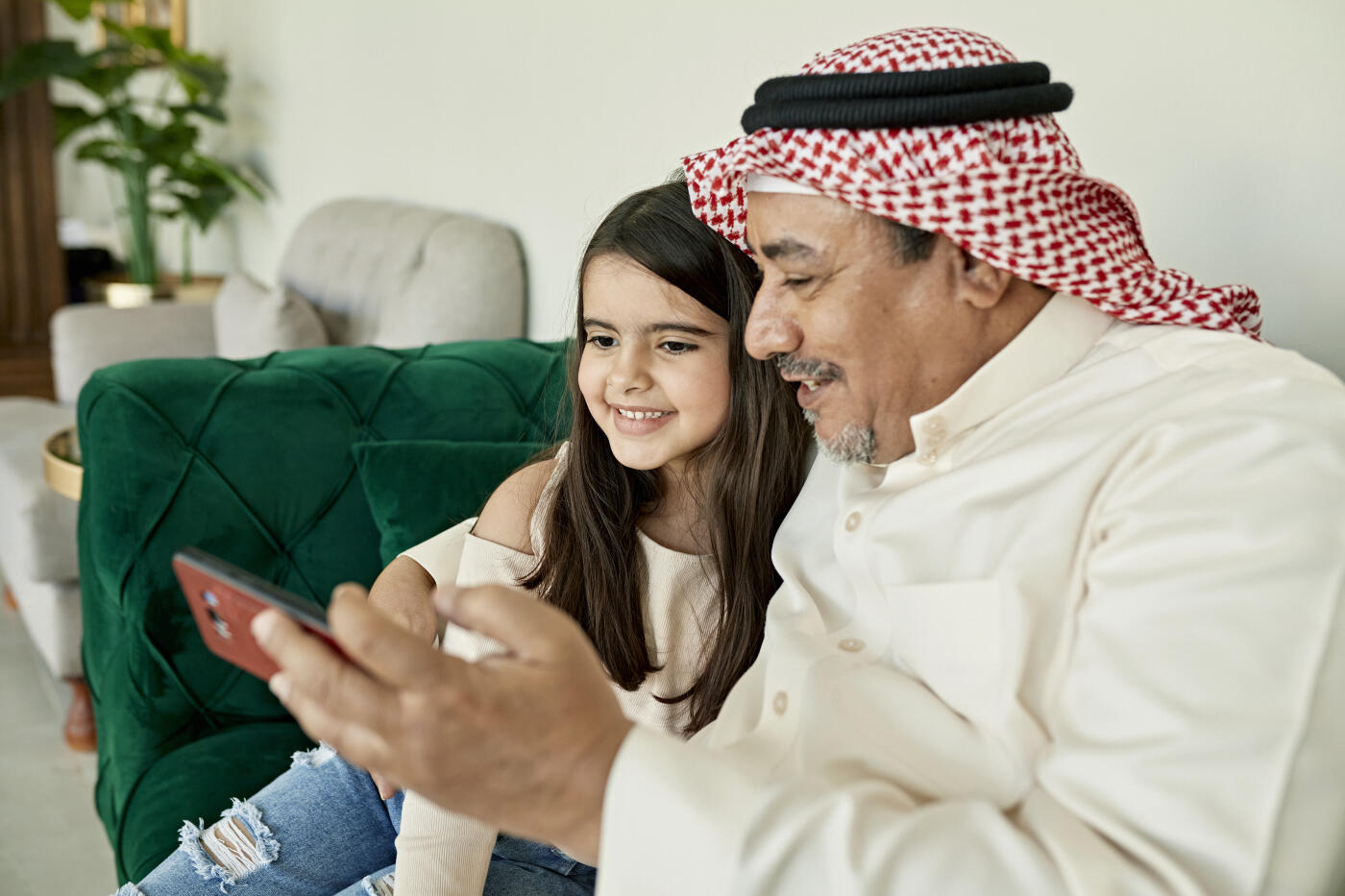 Middle Eastern man in traditional attire with arm around child in casual western clothing,  sitting together on sofa and smiling at smart phone.