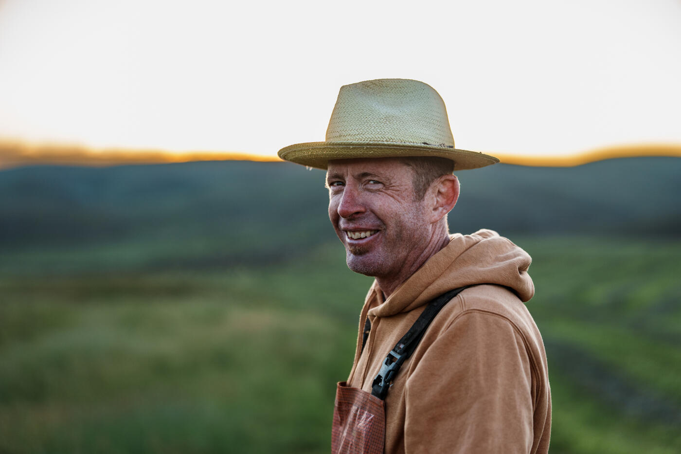 A cheerful rancher wearing waterproof bibs smiles while standing in a dew-covered hay field on a peaceful Wyoming morning.