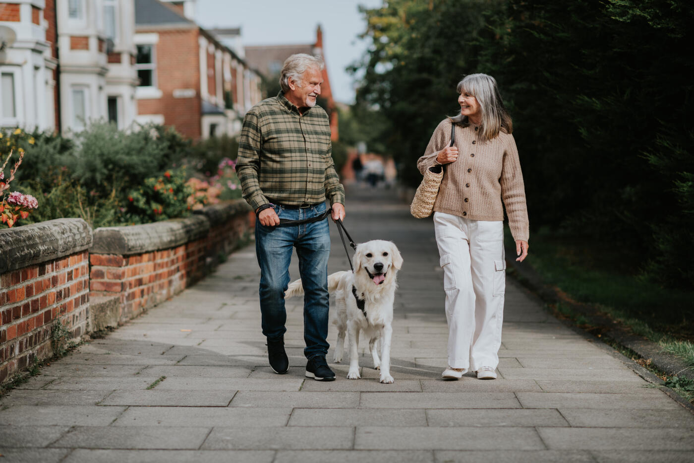 A couple stands on a paved walkway in a park, happily holding their golden retriever dog on a leash. The woman carries a woven bag, and both wear casual outfits.