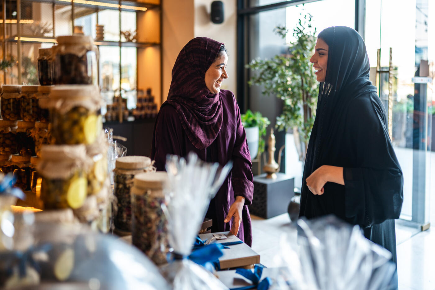 Young Arab women in upscale café engaging with gourmet gift items, smiling and conversing with each other while standing near a display of jars and wrapped goods, surrounded by modern décor and natural light.