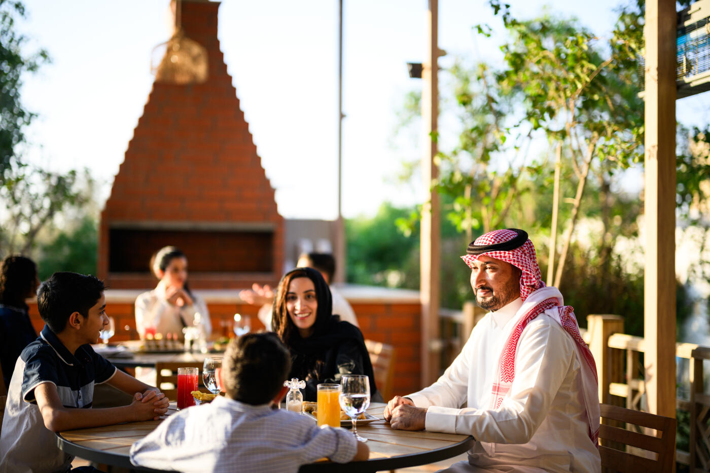 Arab parents and their two sons sitting together at a wooden table on an outdoor restaurant patio. The family enjoying refreshing drinks and engaging in happy conversation during the bright afternoon sun. Warm sunlight filtering through the surrounding green trees creating a relaxed, golden atmosphere.