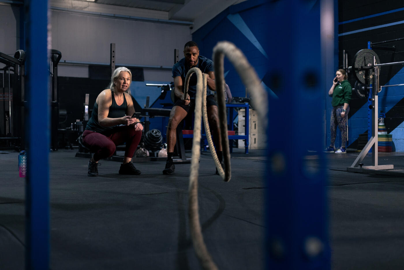 A personal trainer and a mature woman share encouragement at a gym. The woman is dressed in athletic wear, while the trainer, wearing a t-shirt and shorts, shows the client how to perform battle ropes properly. The gym environment features weightlifting equipment and barbells in the background. Located in Newcastle Upon Tyne

Video are available for this scenario