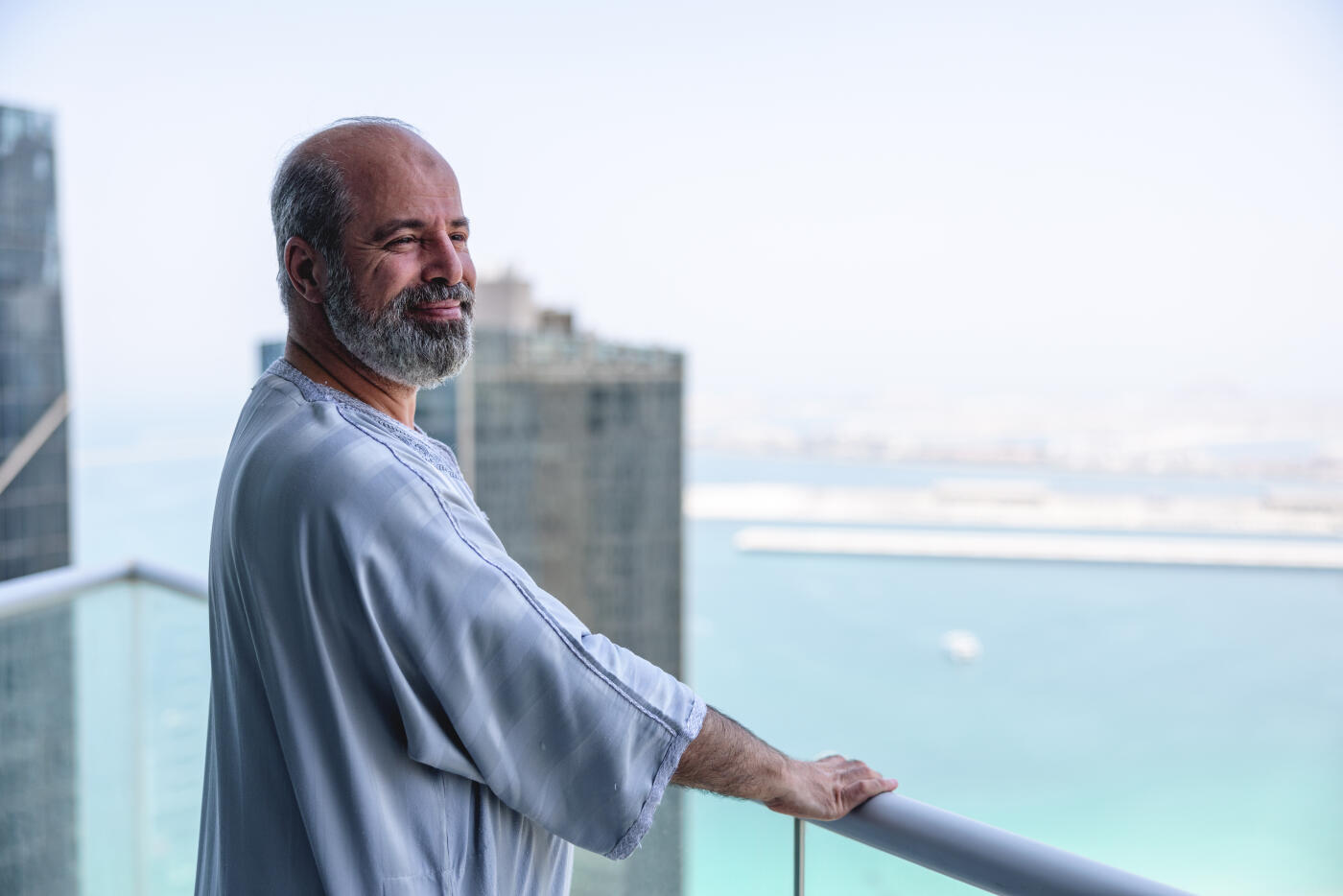 Middle eastern older male is wearing traditional clothes and enjoying beautiful view on his balcony. Large buildings in the background.