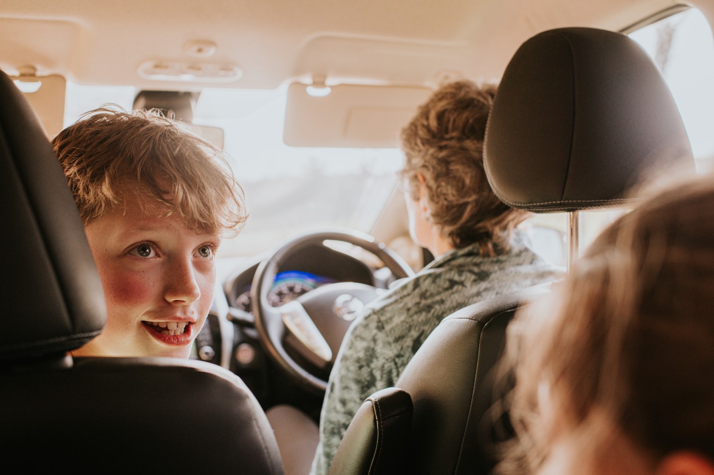 A child sits in the front passenger seat of a right hand drive domestic car. He peers over the arm rest, smiling at the passengers in the back seat.