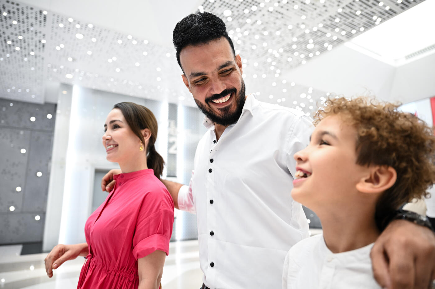 A happy Middle Eastern family, including a woman in a pink dress and a young child, exploring the bright, spacious interior of a modern luxury mall in Dubai. The father is standing close by, interacting with his family as they take in the elegant surroundings.