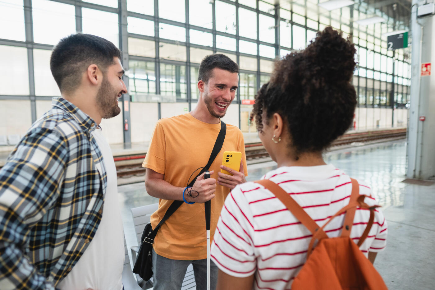 Blind person is standing on a train platform, holding a smartphone and talking with friends