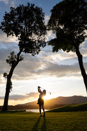 Young hispanic woman swinging golf club on a lush course at sunset, silhouette against golden sky, relaxed and focused recreational golfer