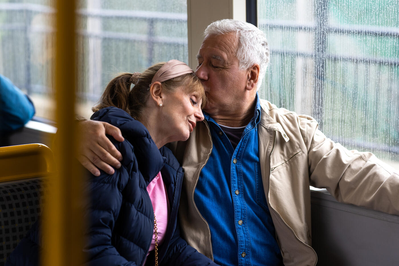 Medium shot of a senior couple sitting side by side on a metro train in North East England. The female is leaning on her partner's shoulder while he has his arm around her shoulder and kisses her on the forehead affectionately.Videos similar to this scenario available.