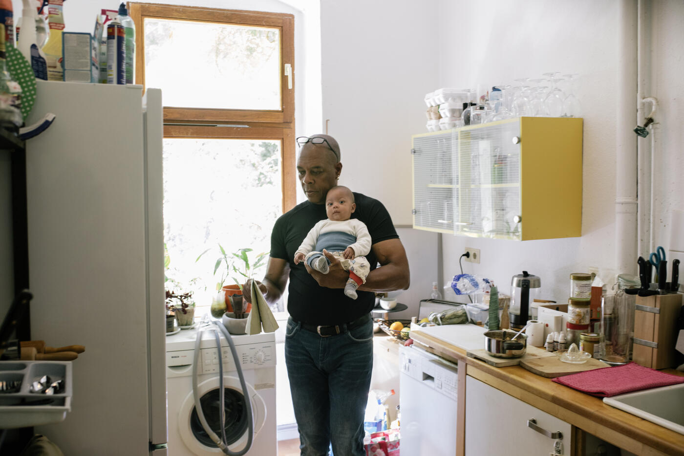 A dad getting ready for lunch at home in his kitchen while holding a young baby in his arms.