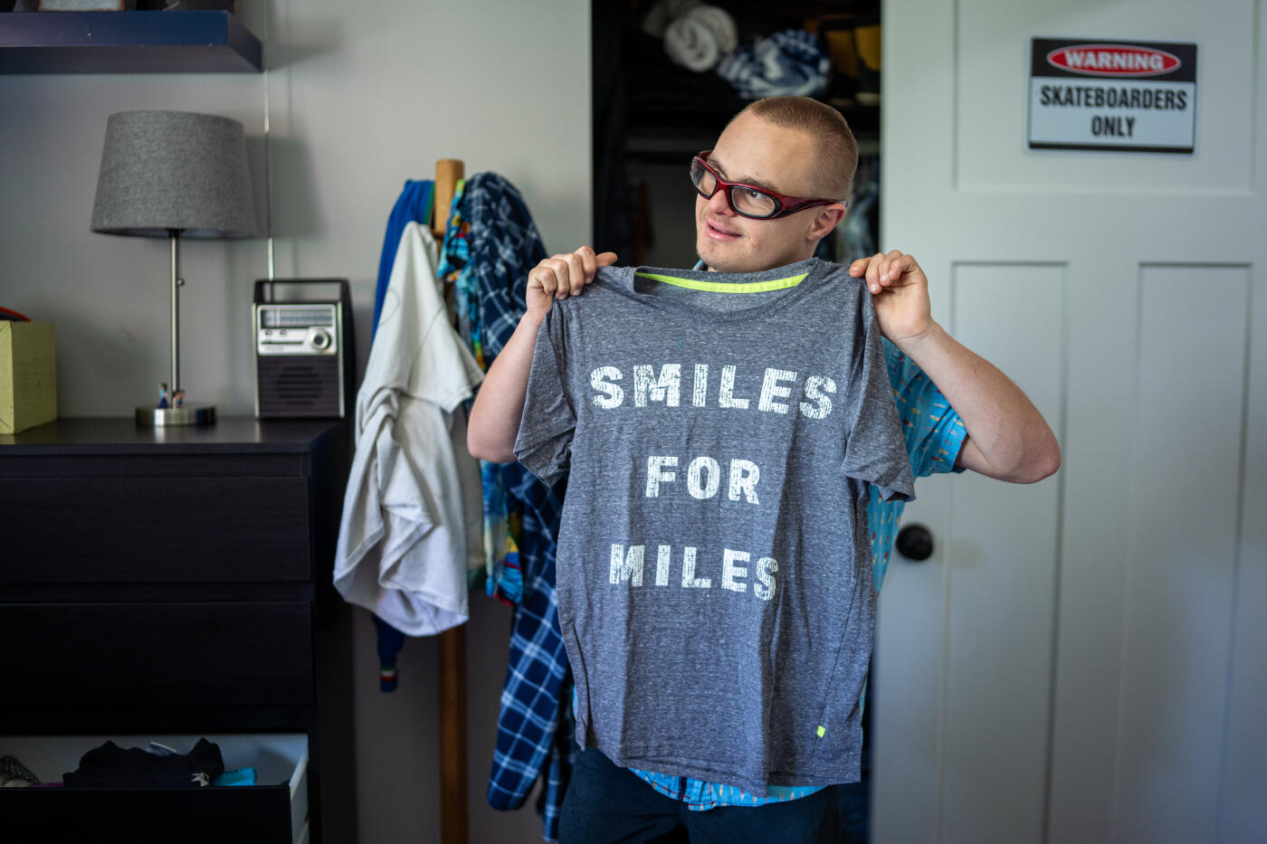 A teenage boy with Down syndrome stands in his bedroom as he holds up a gray T-shirt that reads “Smiles for Miles,” proudly showing off his positive attitude and sense of humor.