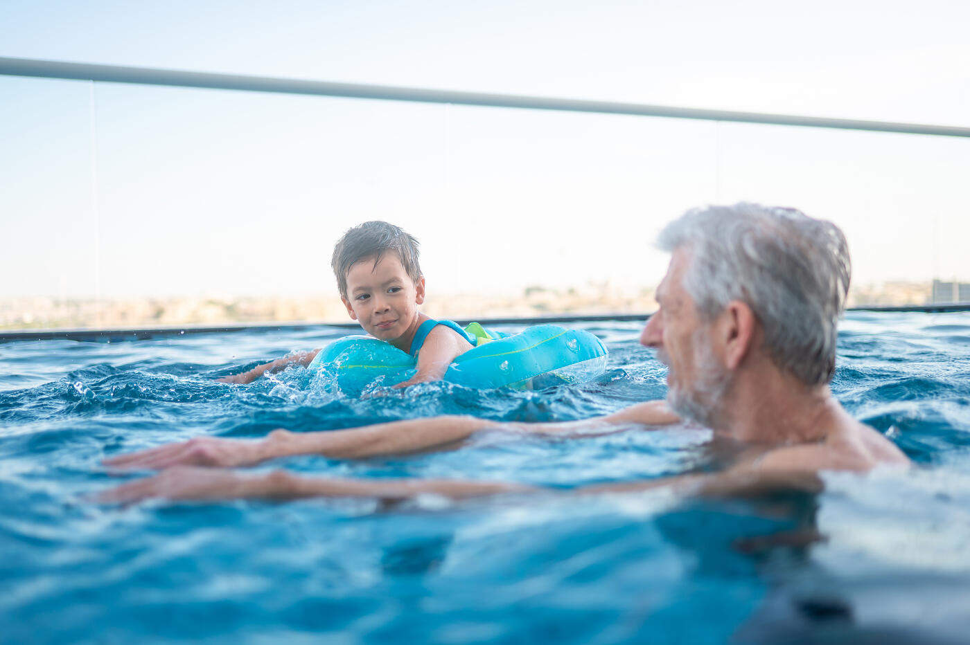 Captured in this image, an elderly man interacts playfully with a young boy in an infinity pool, symbolizing the cherished moments of family bonding during leisure time in the water.