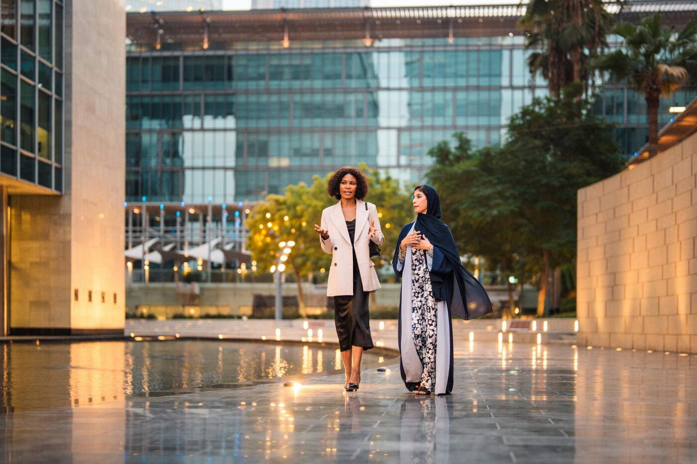 Diverse businesswomen engaging in conversation while walking through the modern architecture of Dubai's financial district. Evening lighting enhances the urban setting, highlighting the professional atmosphere and contemporary design.