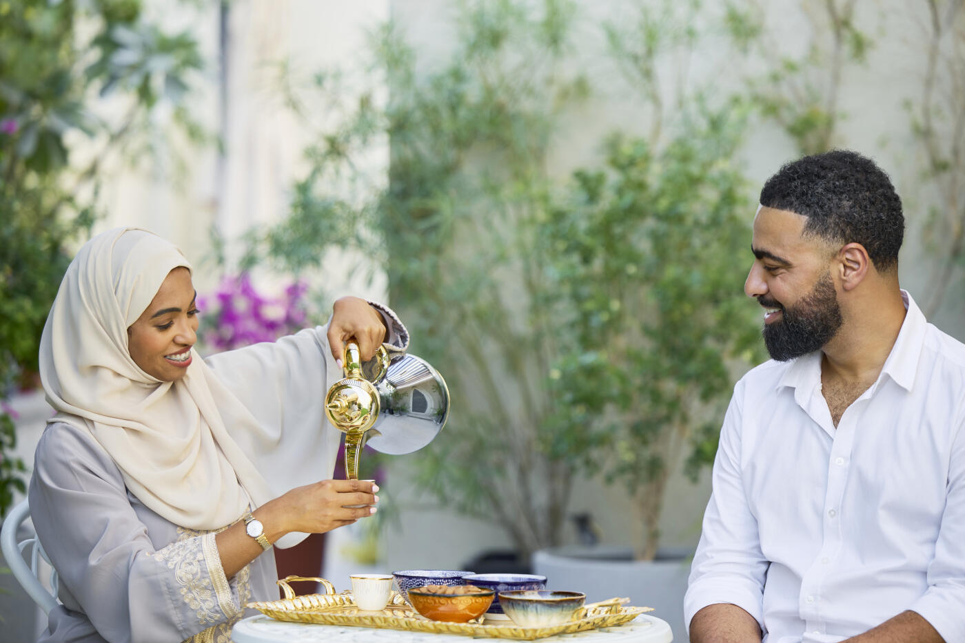 Smiling woman pouring tea while sitting with man in back yard. Saudi female is serving drink. She is wearing hijab.