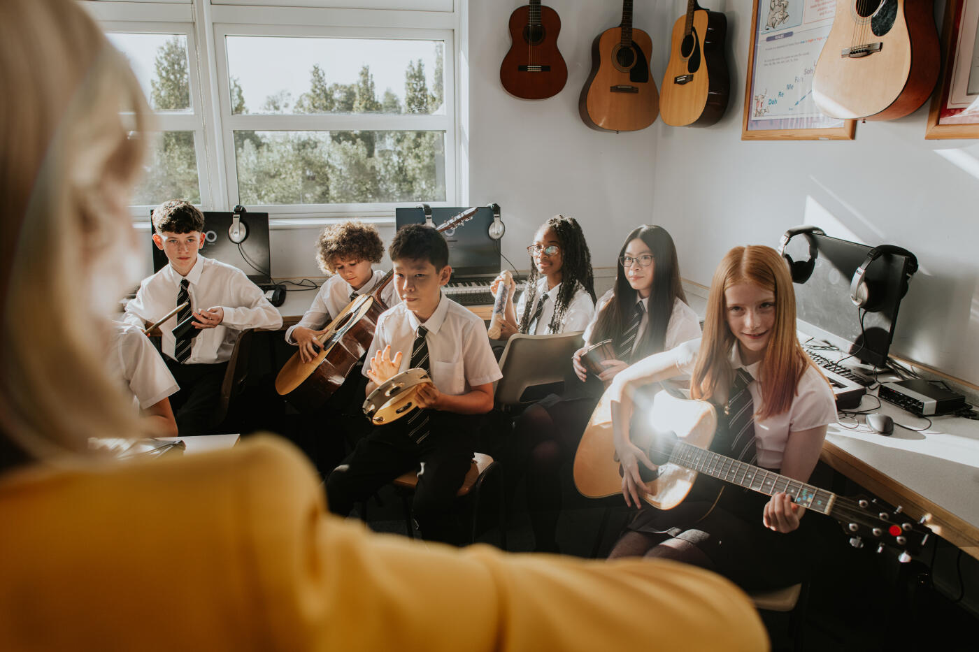Students learn to play various musical instruments in a bright classroom. They are engaged and focused during the lesson, sitting in front of their teacher.