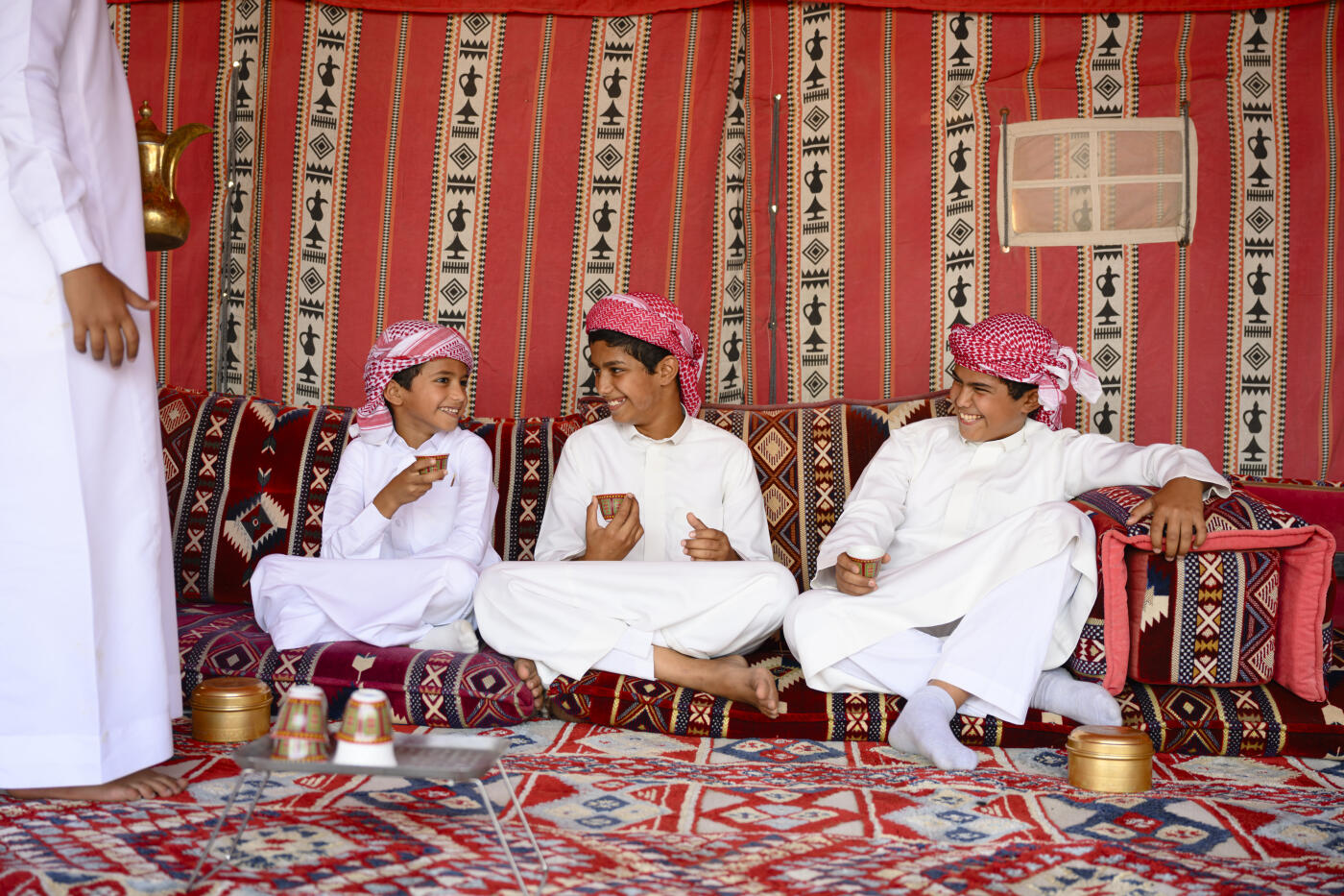 Young men in thobes and ghutras sit on cushions inside a traditional majlis tent, smiling and sharing tea from small cups in a warm, sociable Arabian gathering