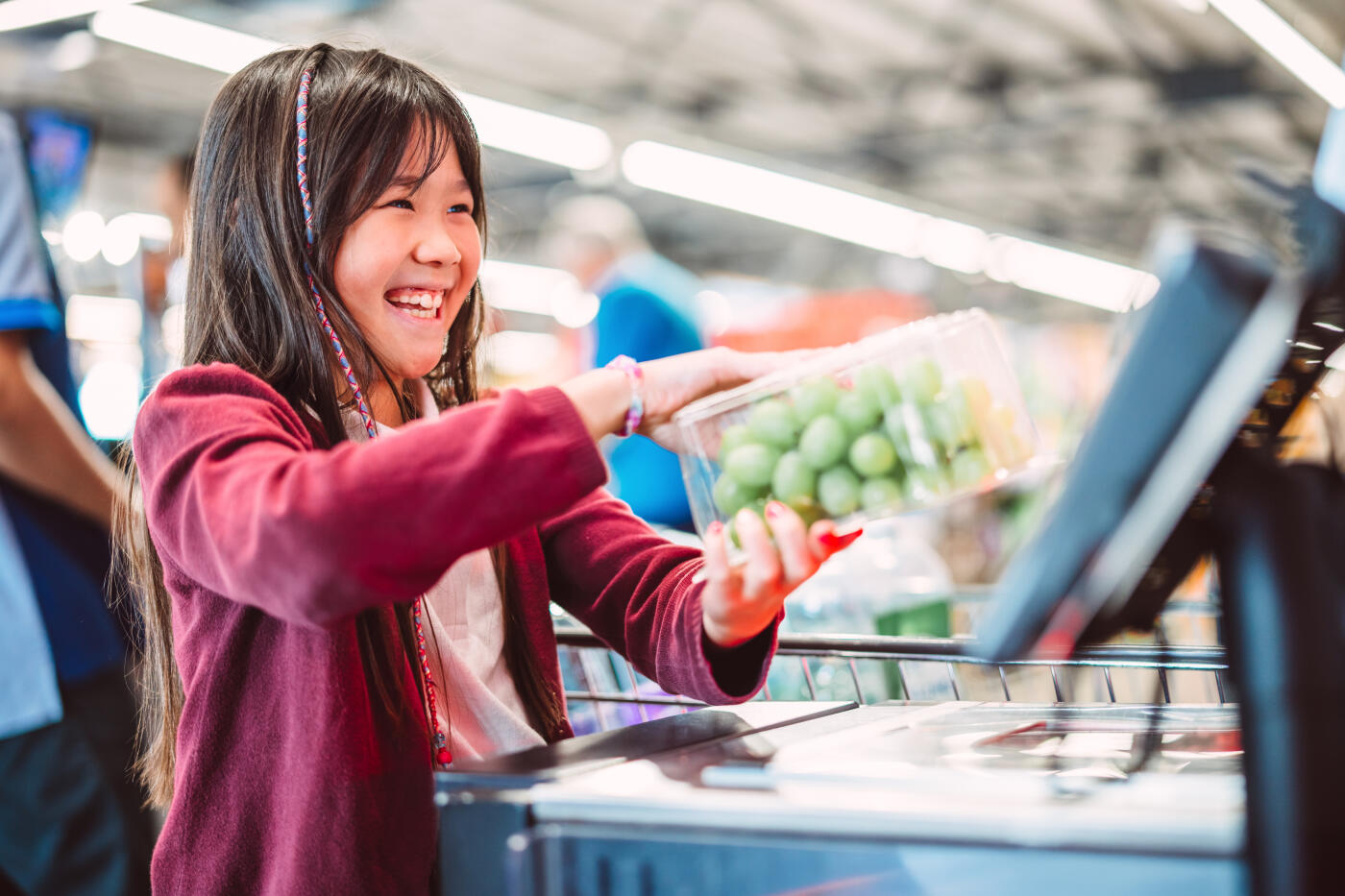 A cheerful young girl scans a pack of fresh green grapes at a self-checkout kiosk in a supermarket. Concepts of family grocery shopping, healthy food choice, sustainable lifestyle, organic produce, and cashless transaction.