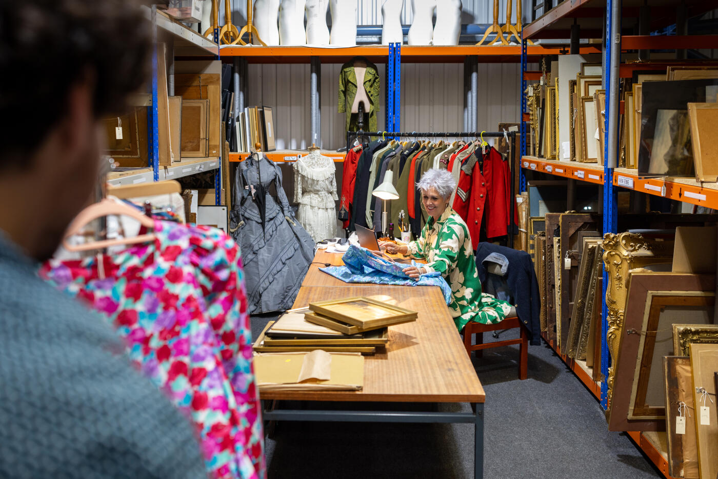 A wide over-the-shoulder shot behind the scenes at an auction house, where a young curator examines a rack of vintage clothing, while a senior colleague works on a laptop at a desk covered in fabrics. The space is filled with antique frames, period costumes, and collectables. They are wearing smart casual clothing in an auction house in the North East of England.

Videos are available similar to this scenario.