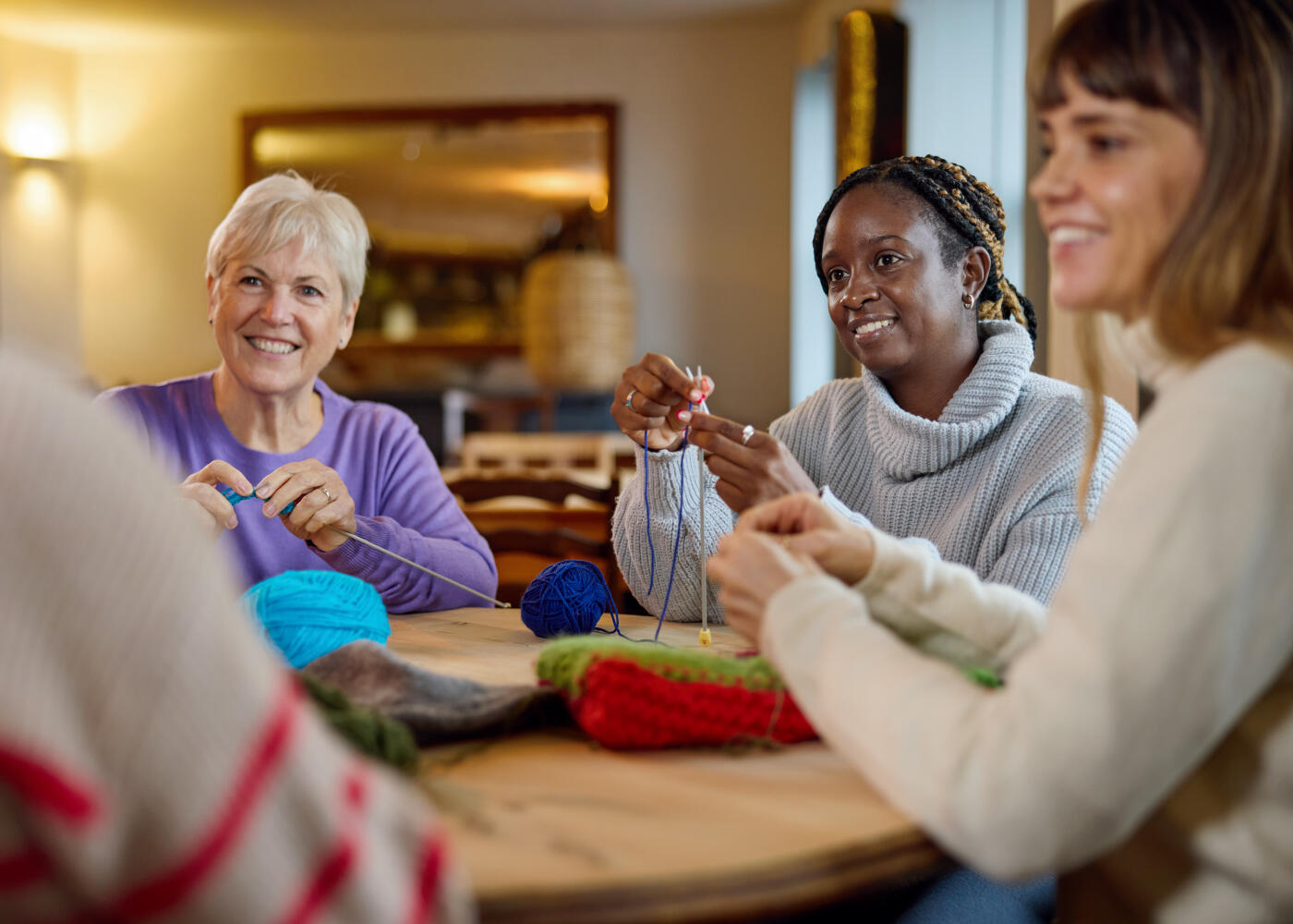 Multiracial group of female friends sitting at wooden table in cafe learning to knit at craft workshop smiling, happiness, friendship, skill