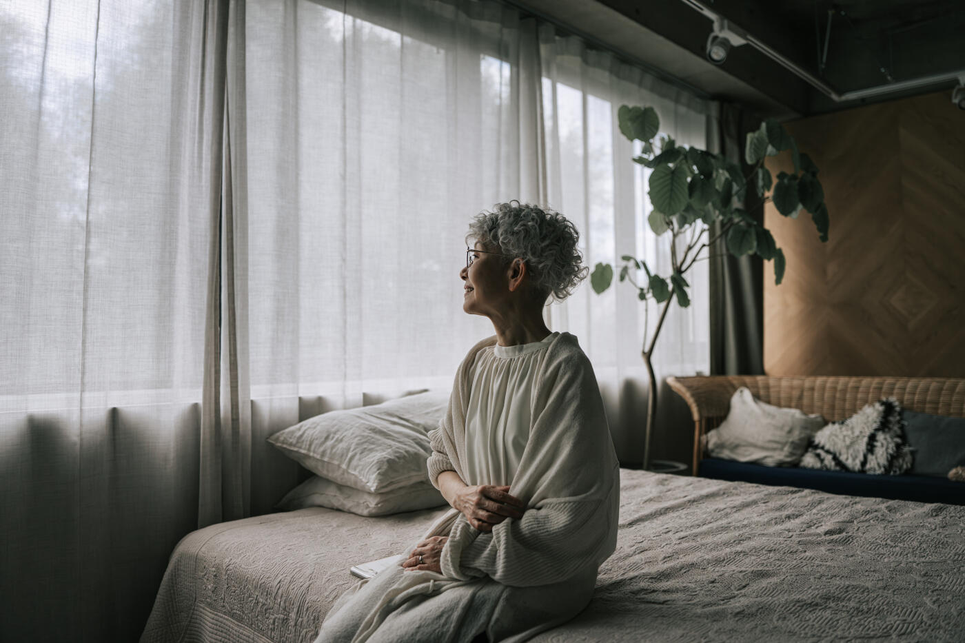 senior Japanese woman with short gray hair and glasses sits calmly on a bed in a cozy, naturally lit bedroom. Dressed in soft, neutral-colored loungewear, she gazes thoughtfully out the window, surrounded by sheer curtains and minimalist decor. The peaceful atmosphere suggests mindfulness, relaxation, healthy aging, and a quiet lifestyle at home.