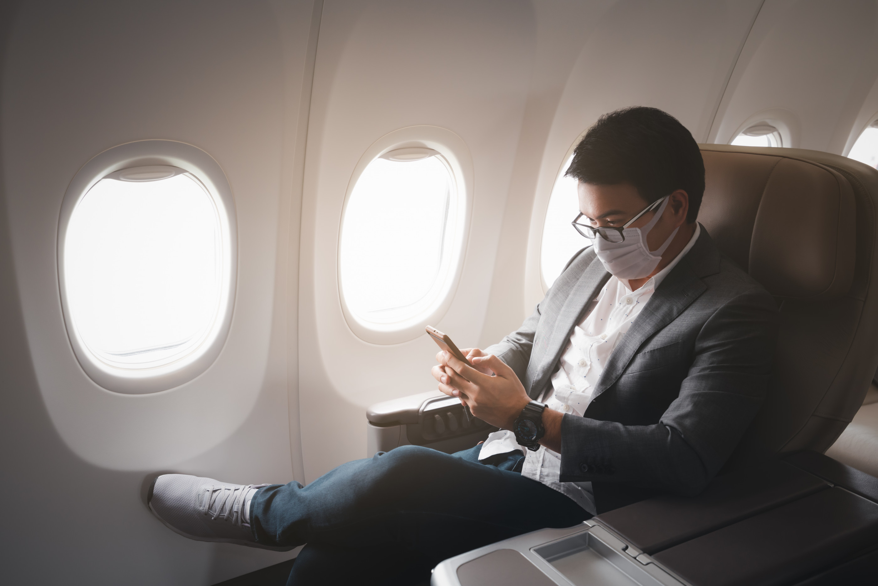 Young asian businessman wearing protective face mask with suit sitting in business first class seat using smartphone due to Coronavirus or COVID-19 outbreak situation in all of landmass in the world