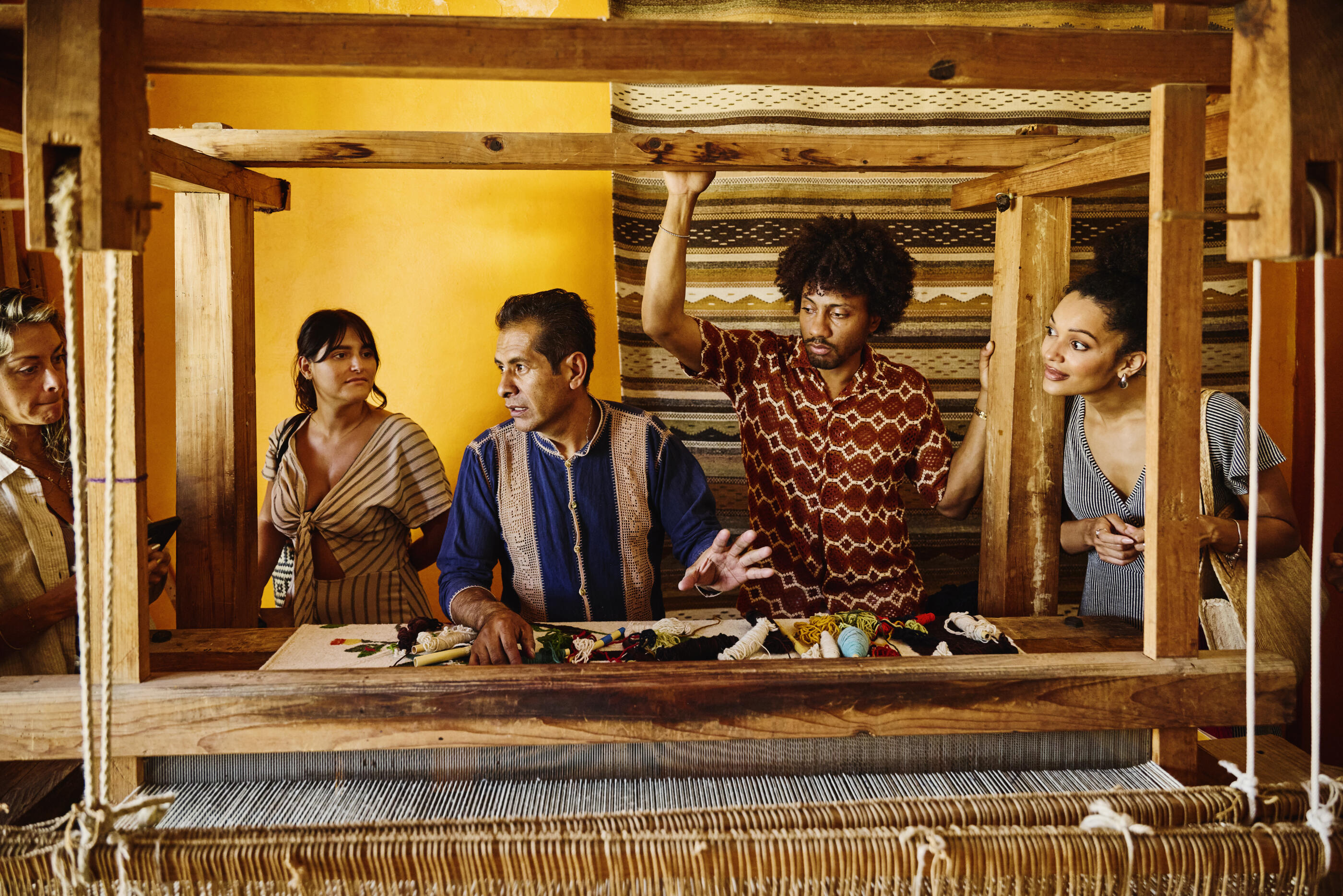 Medium wide shot of male artisan explaining traditional Oaxacan weaving techniques to group of curious tourists during cultural workshop experience