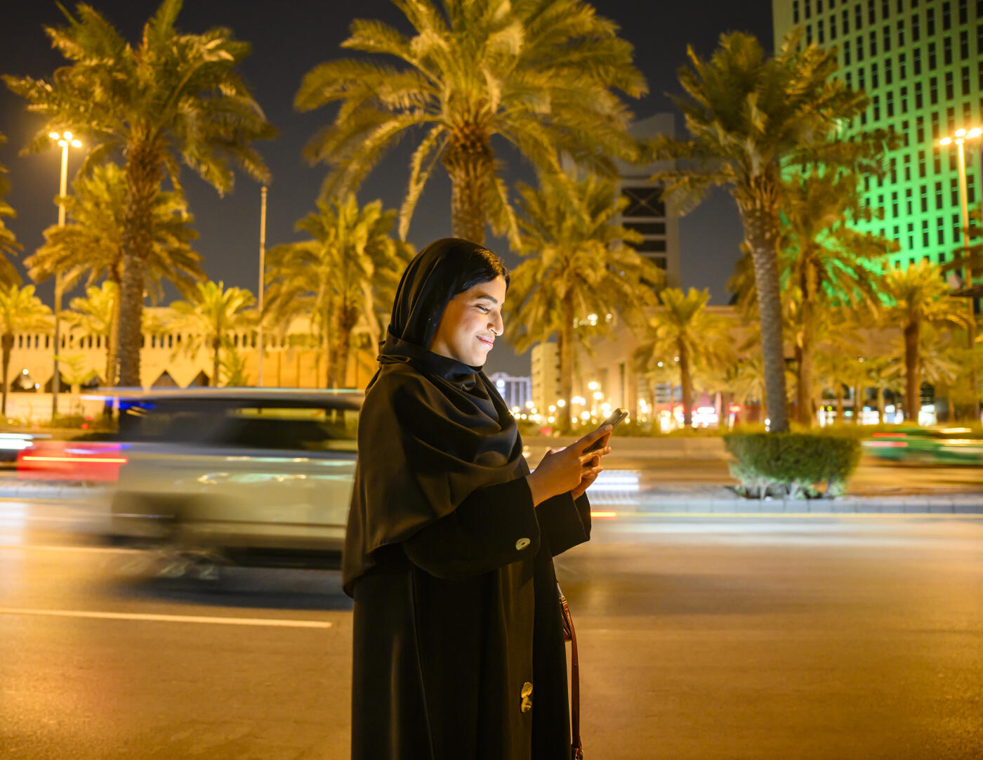 Saudi woman in abaya and hijab smiling while using a smartphone on a lively Riyadh street at night, lit by city lights and light trails, conveying modern urban connectivity and joy