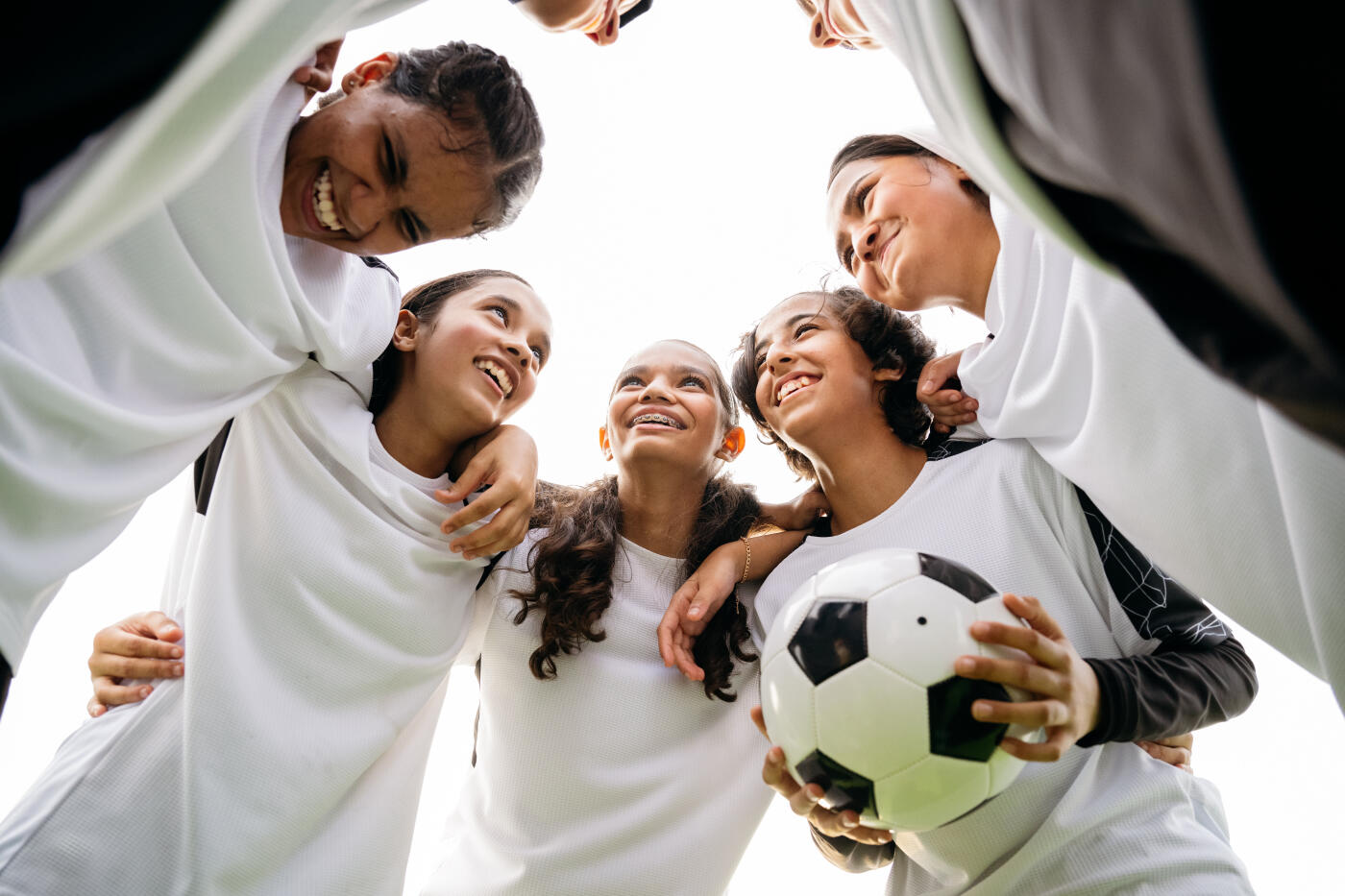Low angle view of cheerful soccer players standing in huddle with ball. Female athletes are with arms around each other. They are planning strategy together.