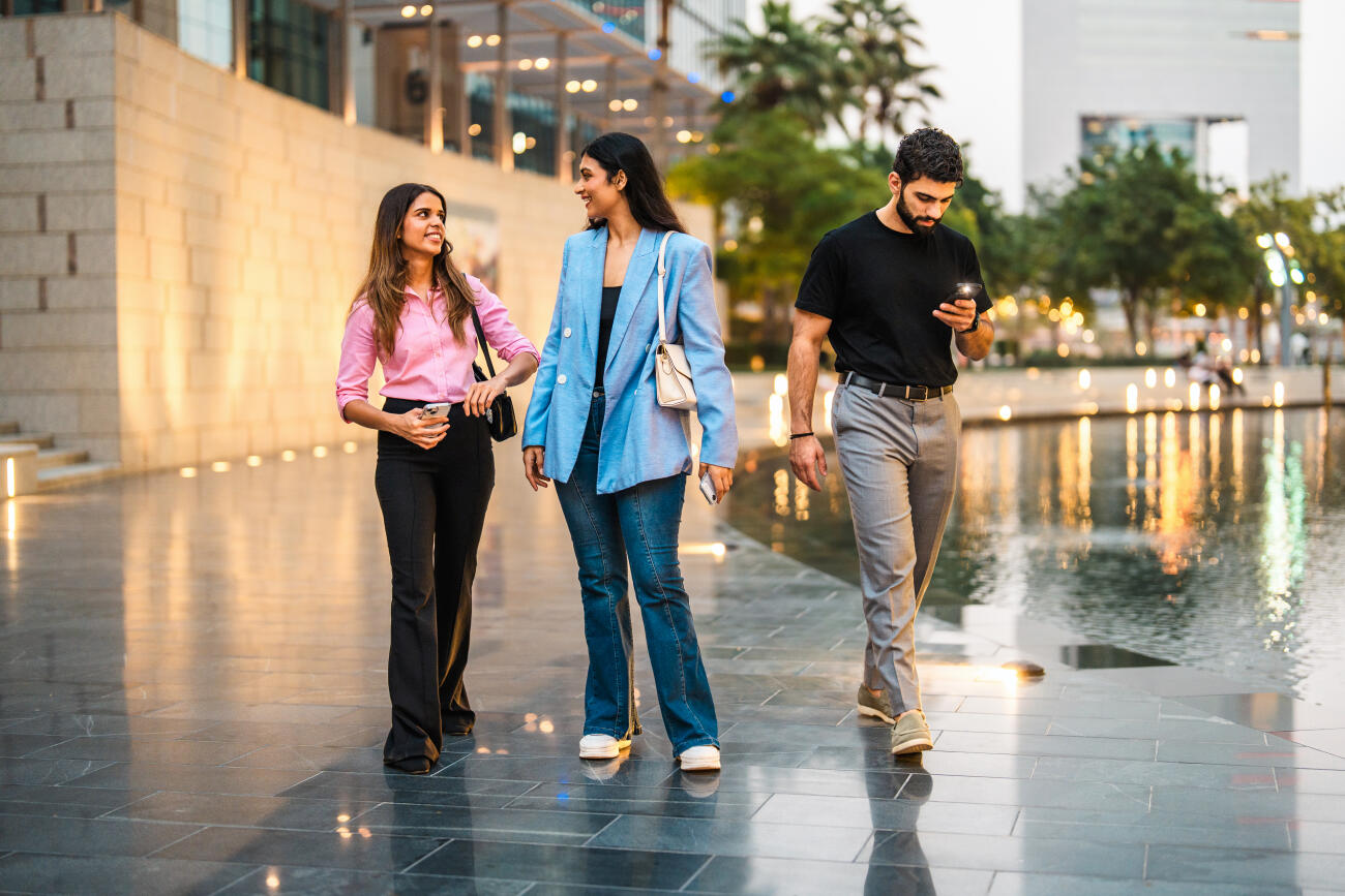 Middle Eastern businesswomen walking along a modern waterfront in Dubai with illuminated buildings in the background. Evening lights reflect on the water, creating a vibrant urban atmosphere.