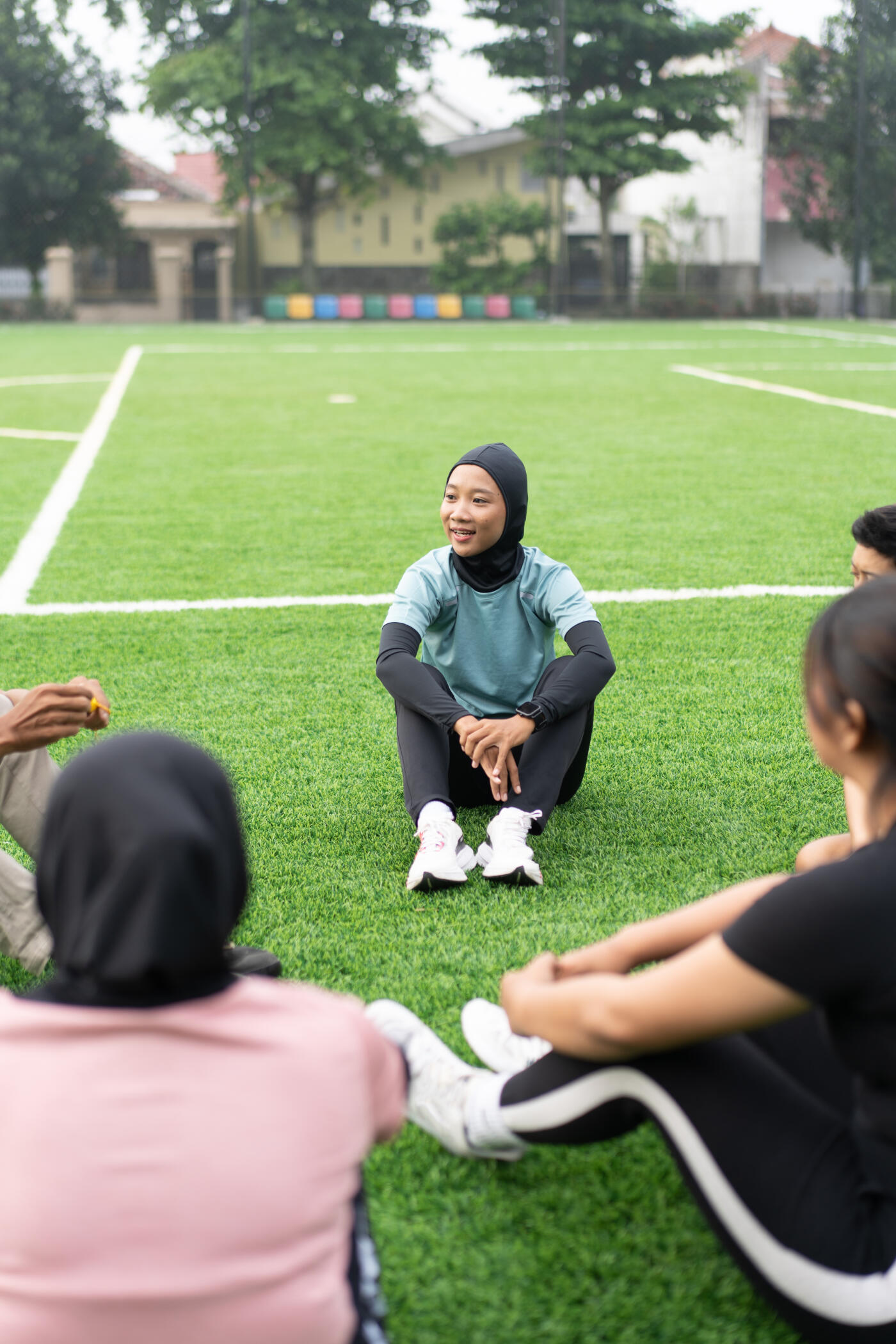 A young female football player wearing a sports hijab and athletic wear sits cross-legged on the artificial grass field, engaging in a discussion with teammates.