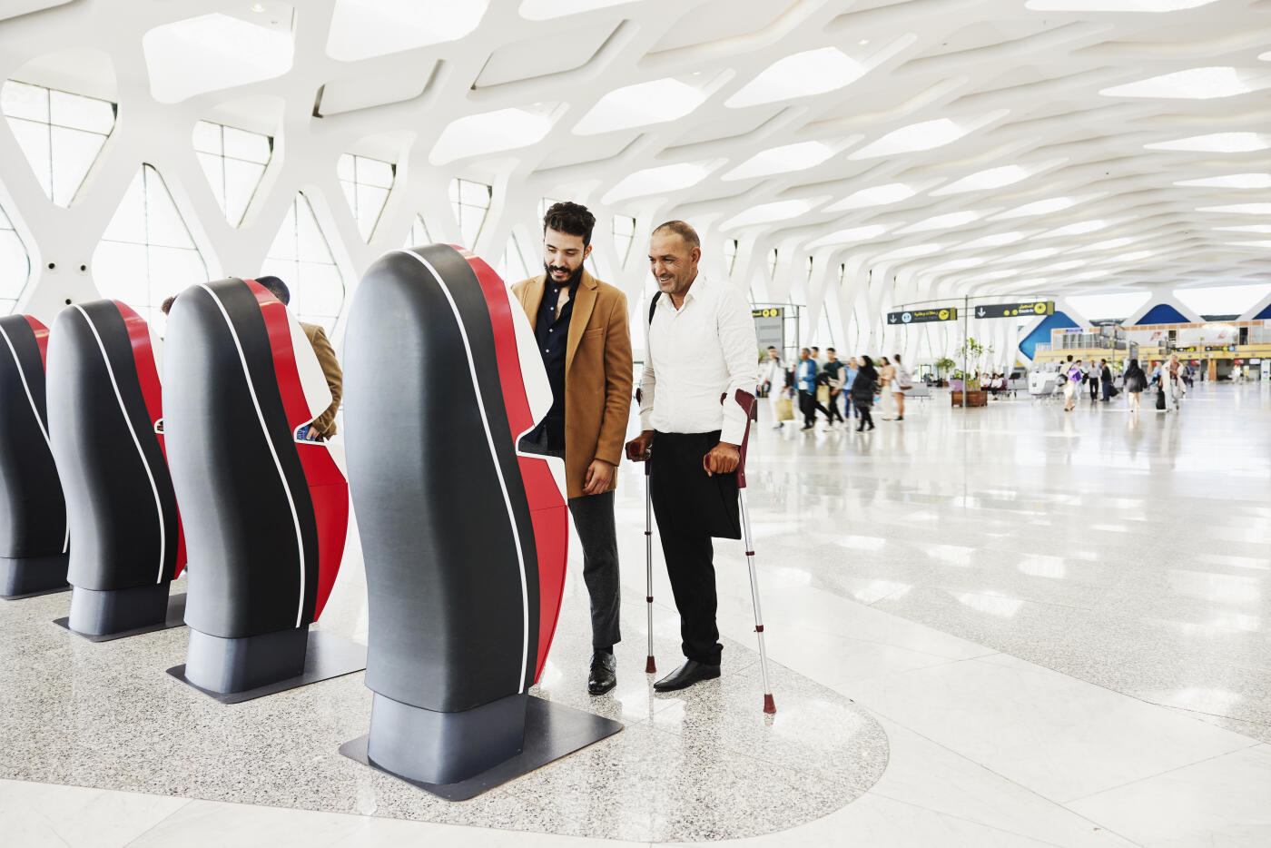 Wide shot of man checking in for flight with disabled friend at electronic self service kiosk in airport terminal